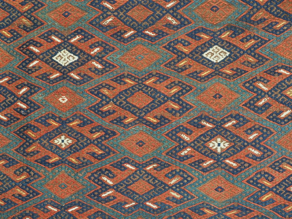 A rectangular handwoven wool kilim rug featuring a dense geometric diamond lattice pattern in terracotta red, navy blue, and cream with wide plain-woven border ends. View X
