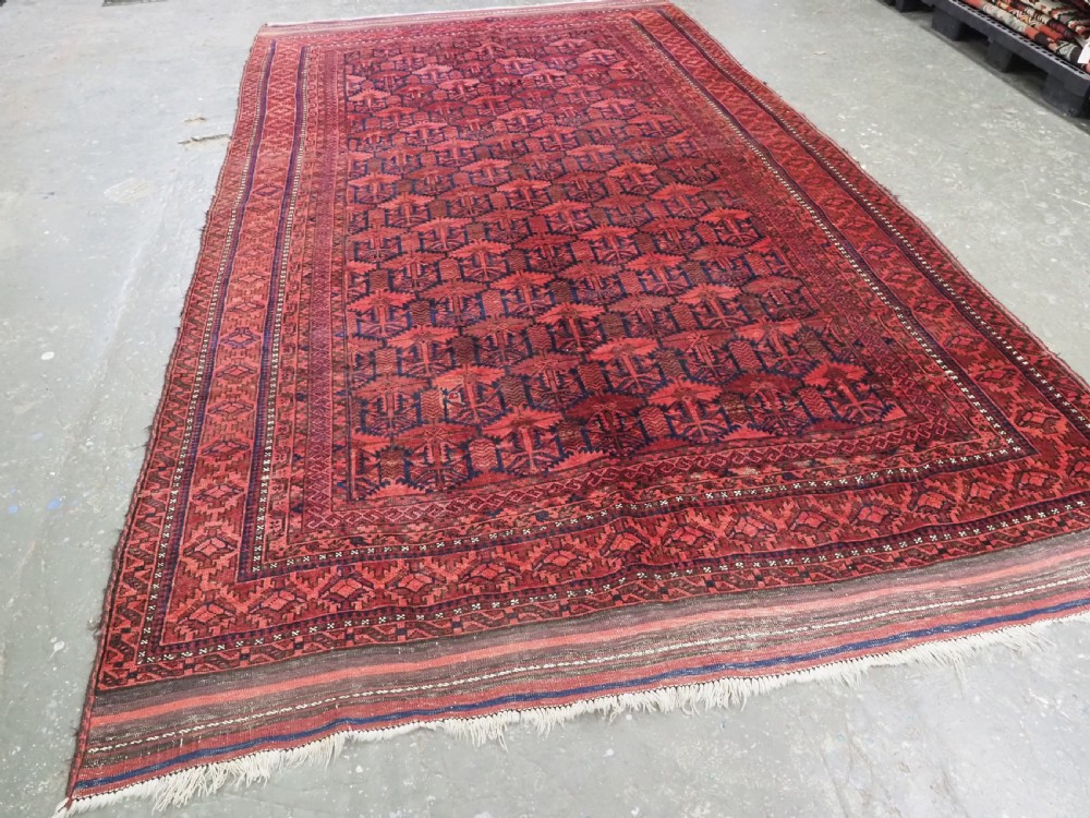 An antique hand-knotted Baluch wool main carpet featuring a deep red field with repeated dark blue geometric motifs and multi-bordered edges. View II