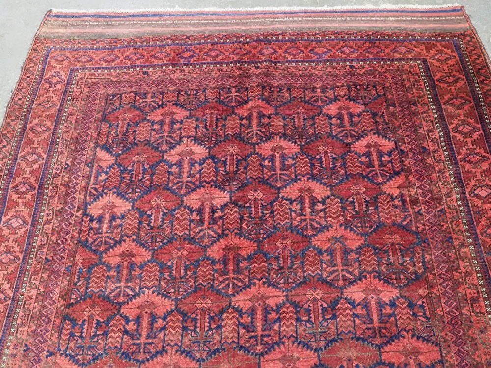 An antique hand-knotted Baluch wool main carpet featuring a deep red field with repeated dark blue geometric motifs and multi-bordered edges. View III