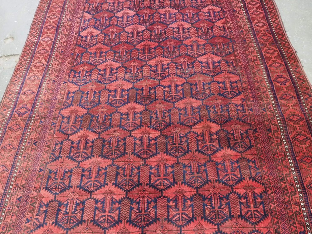 An antique hand-knotted Baluch wool main carpet featuring a deep red field with repeated dark blue geometric motifs and multi-bordered edges. View IV