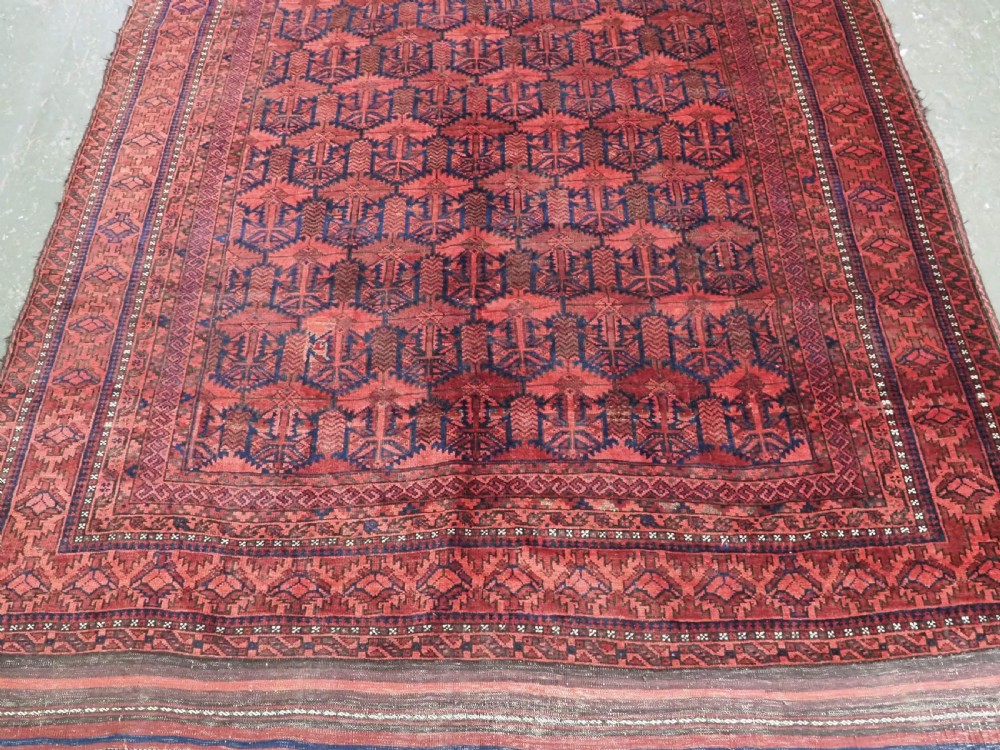 An antique hand-knotted Baluch wool main carpet featuring a deep red field with repeated dark blue geometric motifs and multi-bordered edges. View V