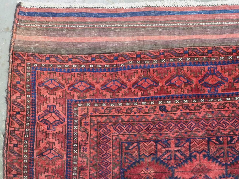 An antique hand-knotted Baluch wool main carpet featuring a deep red field with repeated dark blue geometric motifs and multi-bordered edges. View VI