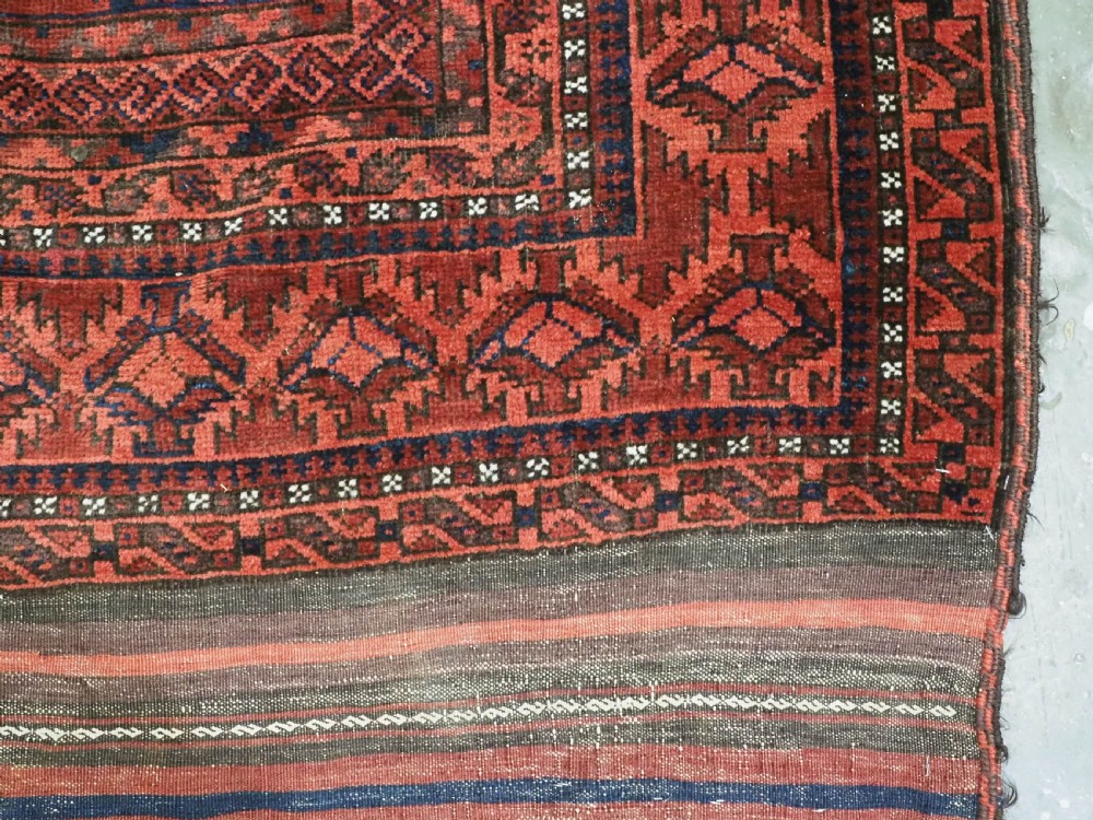 An antique hand-knotted Baluch wool main carpet featuring a deep red field with repeated dark blue geometric motifs and multi-bordered edges. View VII