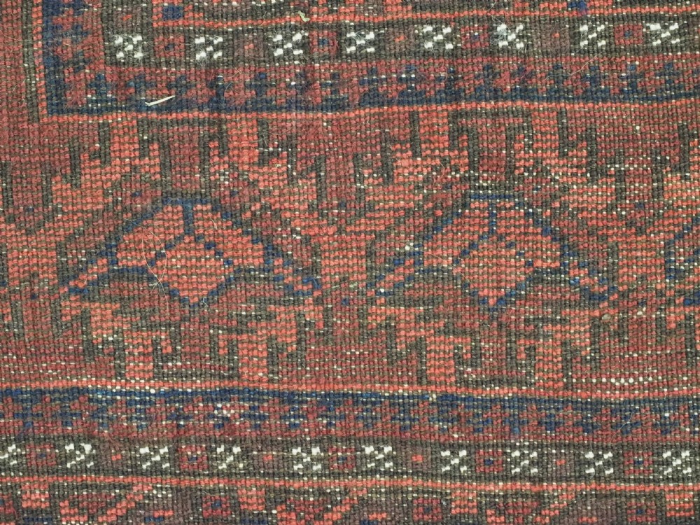 An antique hand-knotted Baluch wool main carpet featuring a deep red field with repeated dark blue geometric motifs and multi-bordered edges. View IX