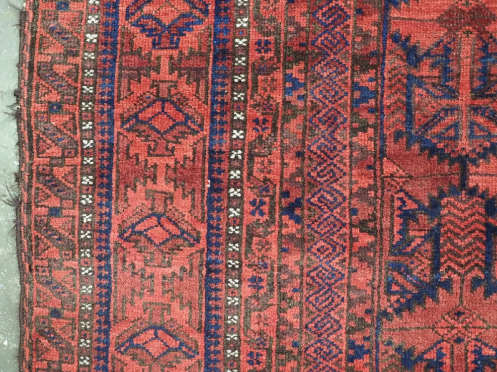 An antique hand-knotted Baluch wool main carpet featuring a deep red field with repeated dark blue geometric motifs and multi-bordered edges. View X