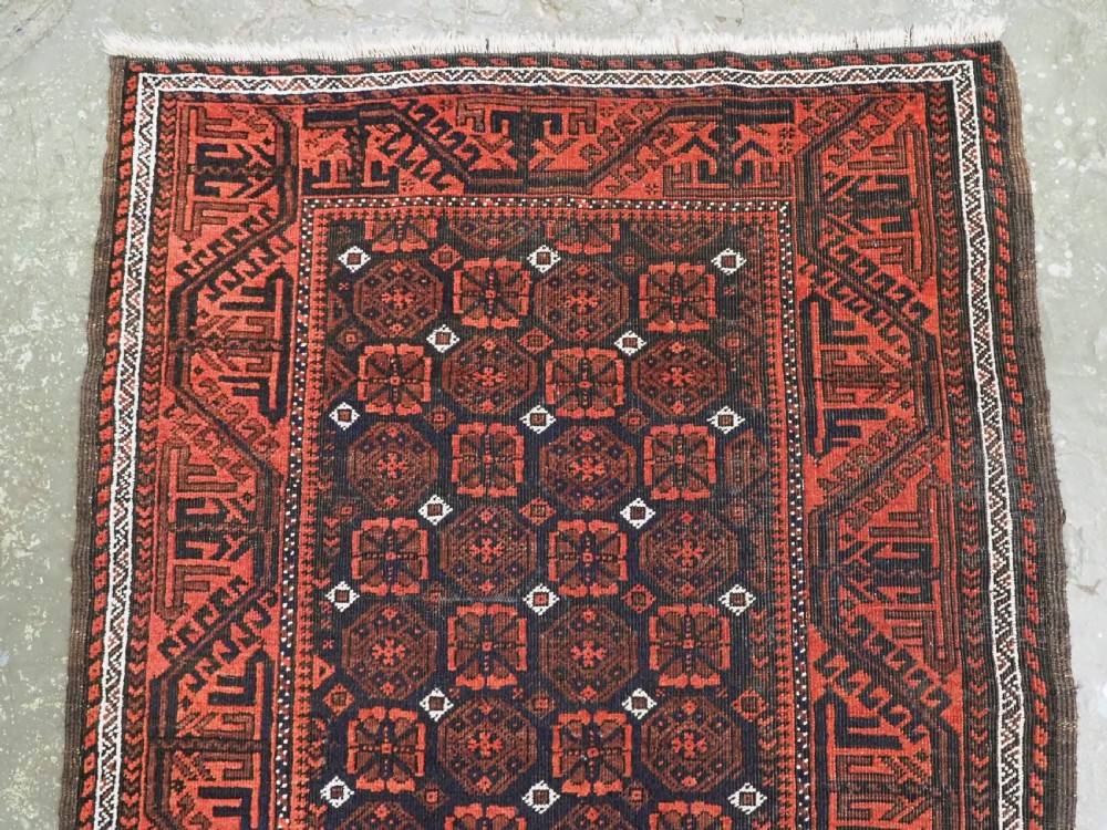 An antique handwoven Baluch rug featuring a dark central field with repeating geometric floral motifs and a wide terracotta red border. View II