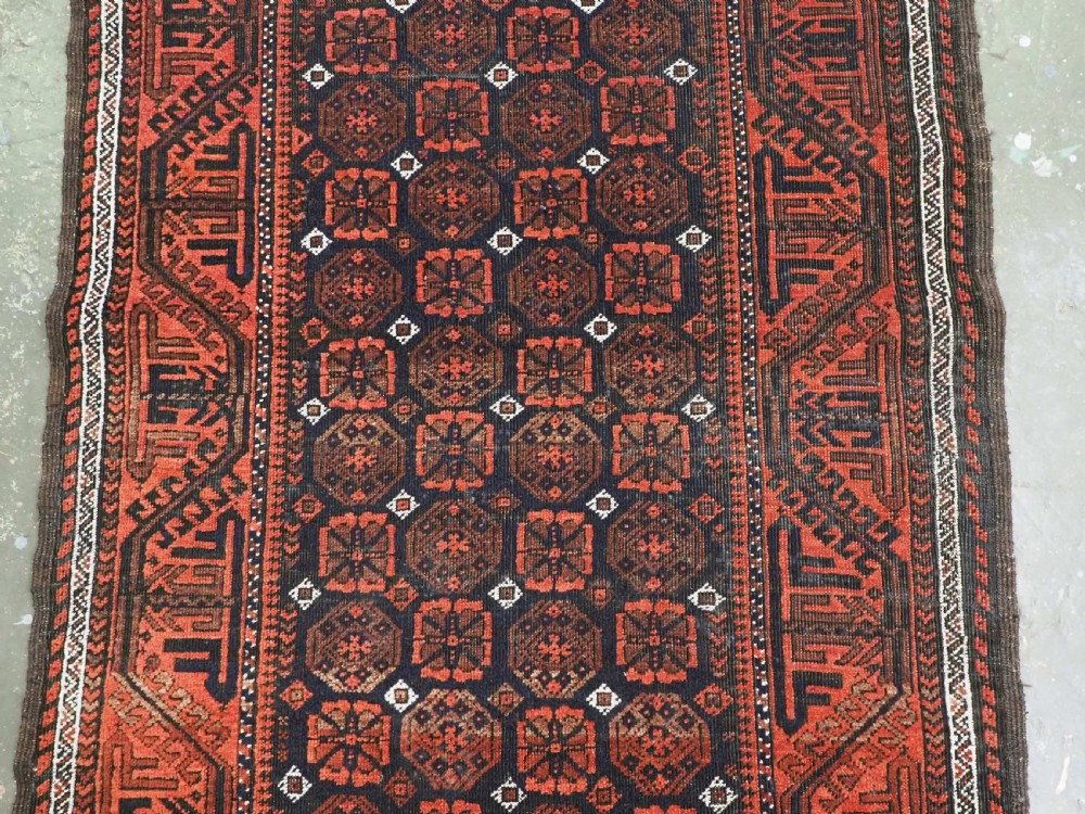 An antique handwoven Baluch rug featuring a dark central field with repeating geometric floral motifs and a wide terracotta red border. View III