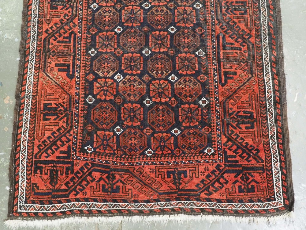 An antique handwoven Baluch rug featuring a dark central field with repeating geometric floral motifs and a wide terracotta red border. View IV