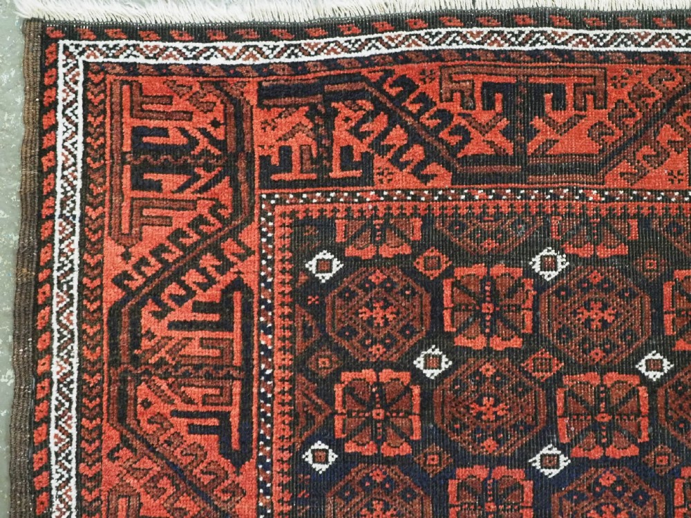An antique handwoven Baluch rug featuring a dark central field with repeating geometric floral motifs and a wide terracotta red border. View V