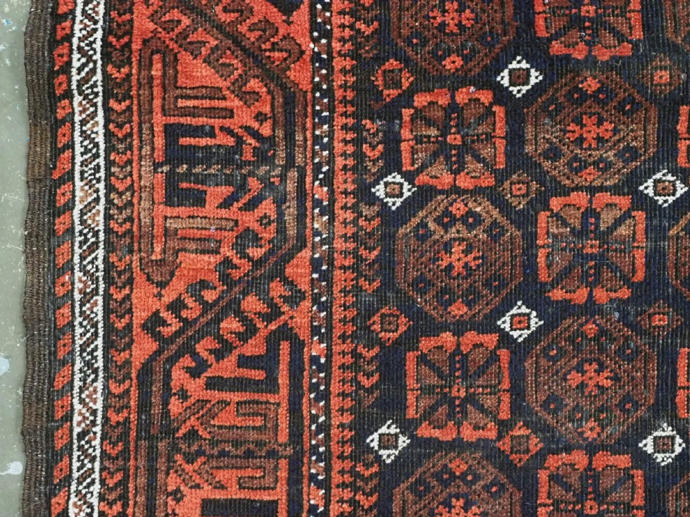 An antique handwoven Baluch rug featuring a dark central field with repeating geometric floral motifs and a wide terracotta red border. View VI