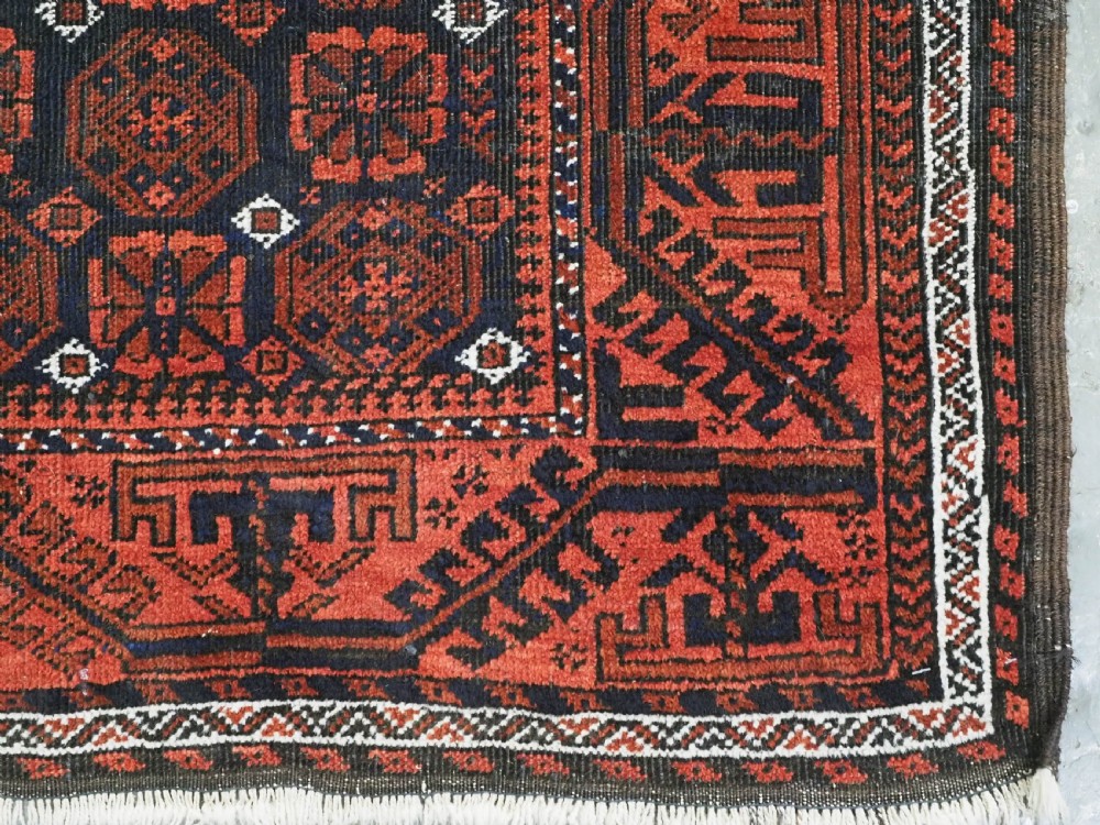 An antique handwoven Baluch rug featuring a dark central field with repeating geometric floral motifs and a wide terracotta red border. View IX