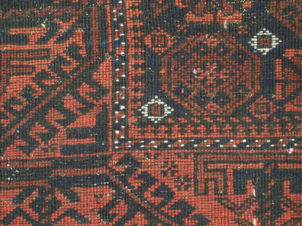 An antique handwoven Baluch rug featuring a dark central field with repeating geometric floral motifs and a wide terracotta red border. View X