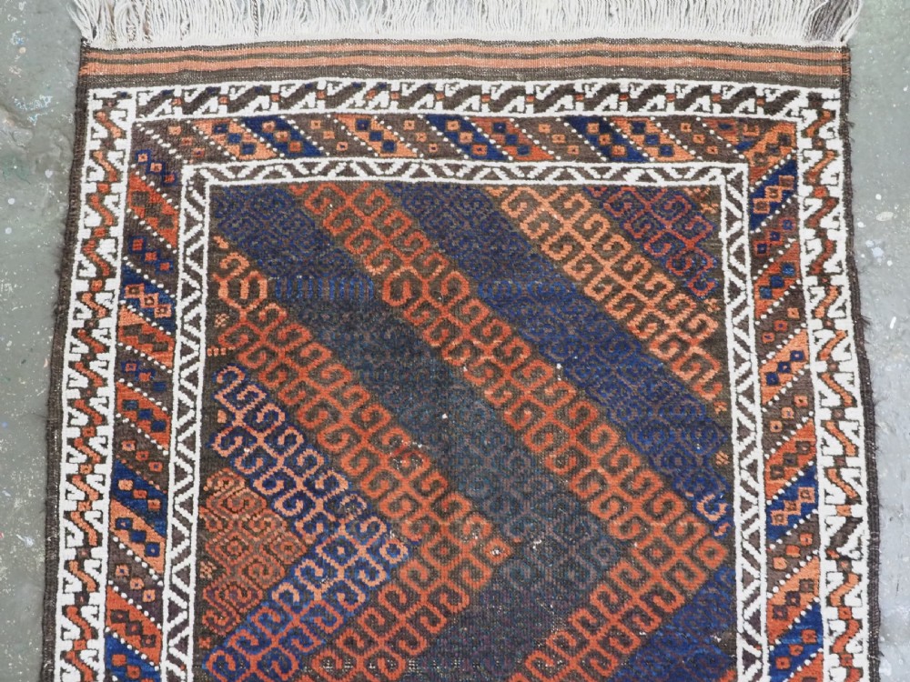 An antique hand-woven Baluch wool rug featuring diagonal geometric chevron patterns in terracotta, indigo blue, and brown surrounded by a cream patterned border. View III