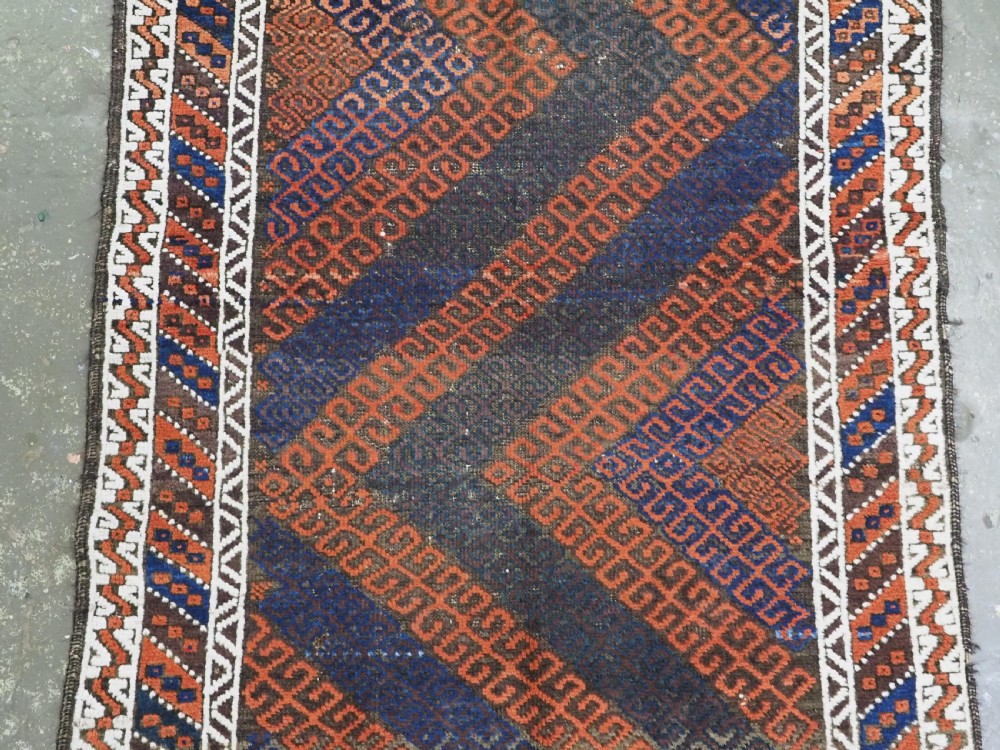 An antique hand-woven Baluch wool rug featuring diagonal geometric chevron patterns in terracotta, indigo blue, and brown surrounded by a cream patterned border. View IV