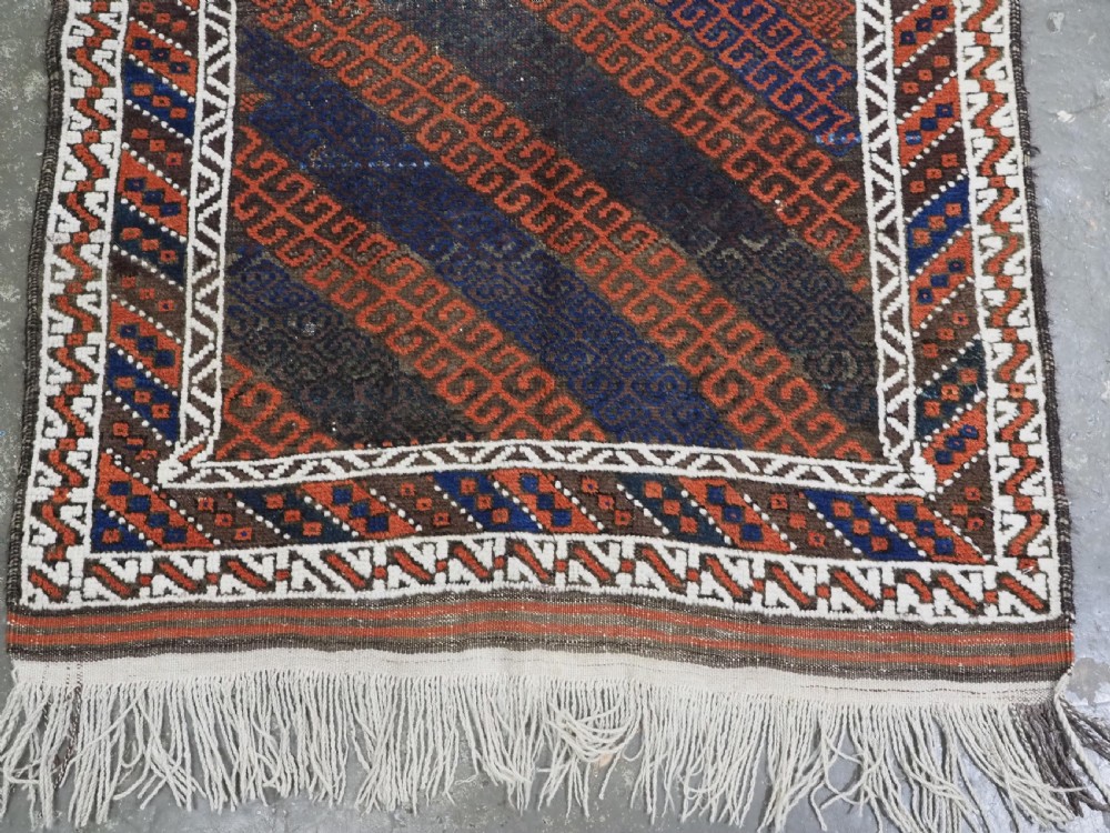 An antique hand-woven Baluch wool rug featuring diagonal geometric chevron patterns in terracotta, indigo blue, and brown surrounded by a cream patterned border. View V