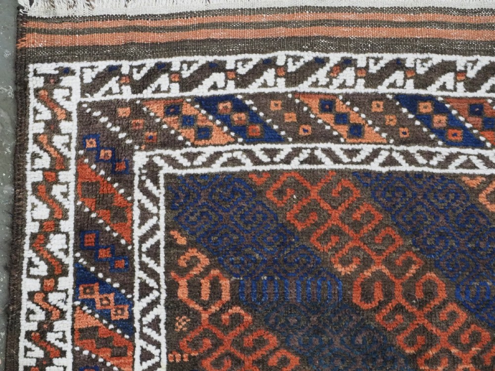 An antique hand-woven Baluch wool rug featuring diagonal geometric chevron patterns in terracotta, indigo blue, and brown surrounded by a cream patterned border. View VI