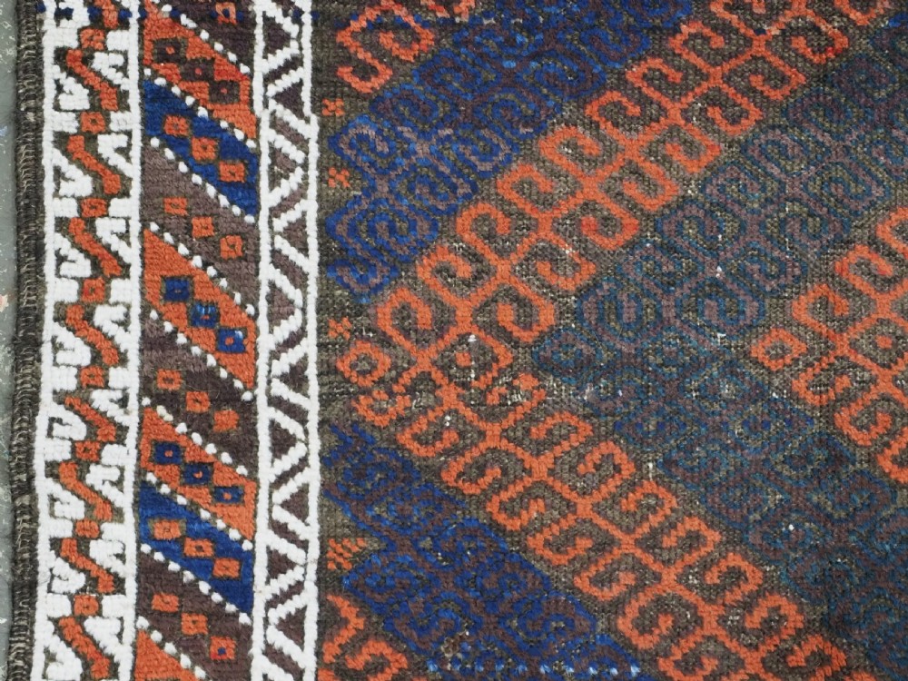 An antique hand-woven Baluch wool rug featuring diagonal geometric chevron patterns in terracotta, indigo blue, and brown surrounded by a cream patterned border. View VII