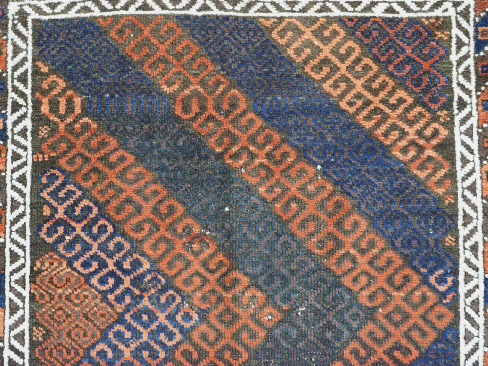 An antique hand-woven Baluch wool rug featuring diagonal geometric chevron patterns in terracotta, indigo blue, and brown surrounded by a cream patterned border. View VIII