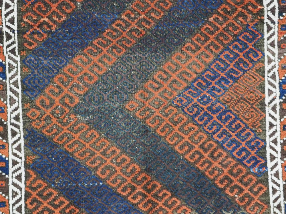 An antique hand-woven Baluch wool rug featuring diagonal geometric chevron patterns in terracotta, indigo blue, and brown surrounded by a cream patterned border. View IX