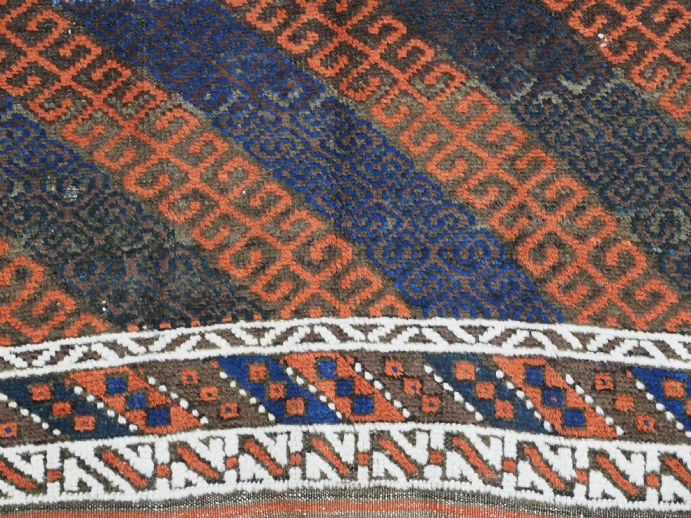 An antique hand-woven Baluch wool rug featuring diagonal geometric chevron patterns in terracotta, indigo blue, and brown surrounded by a cream patterned border. View X