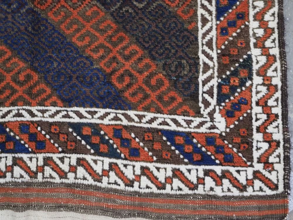 An antique hand-woven Baluch wool rug featuring diagonal geometric chevron patterns in terracotta, indigo blue, and brown surrounded by a cream patterned border. View XI