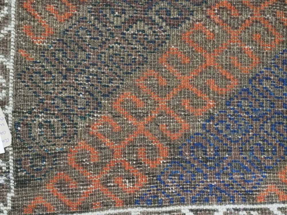 An antique hand-woven Baluch wool rug featuring diagonal geometric chevron patterns in terracotta, indigo blue, and brown surrounded by a cream patterned border. View XII