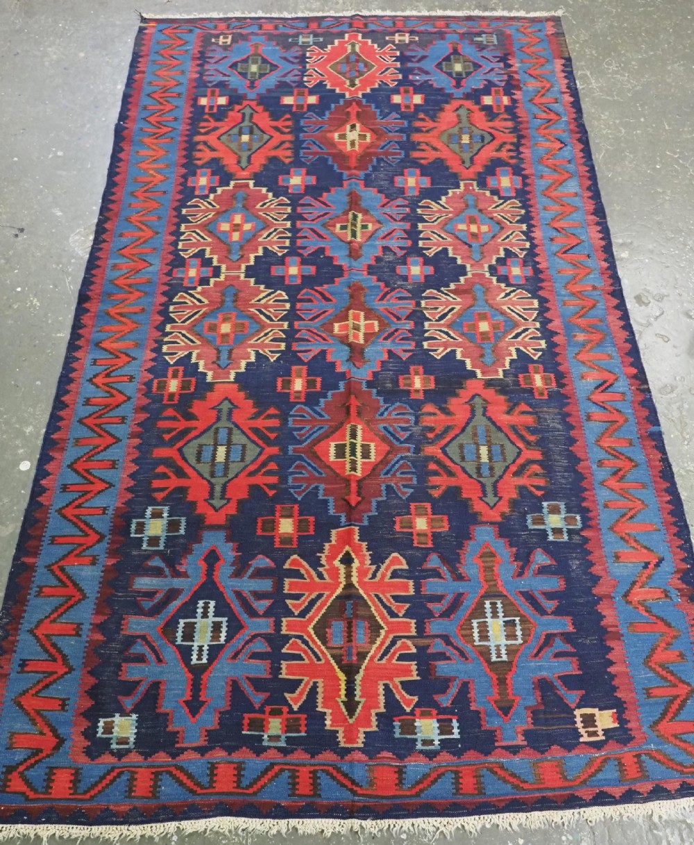 An antique flatweave Caucasian Kuba kilim rug featuring geometric shield medallions in red, blue, and cream across a dark navy field.
