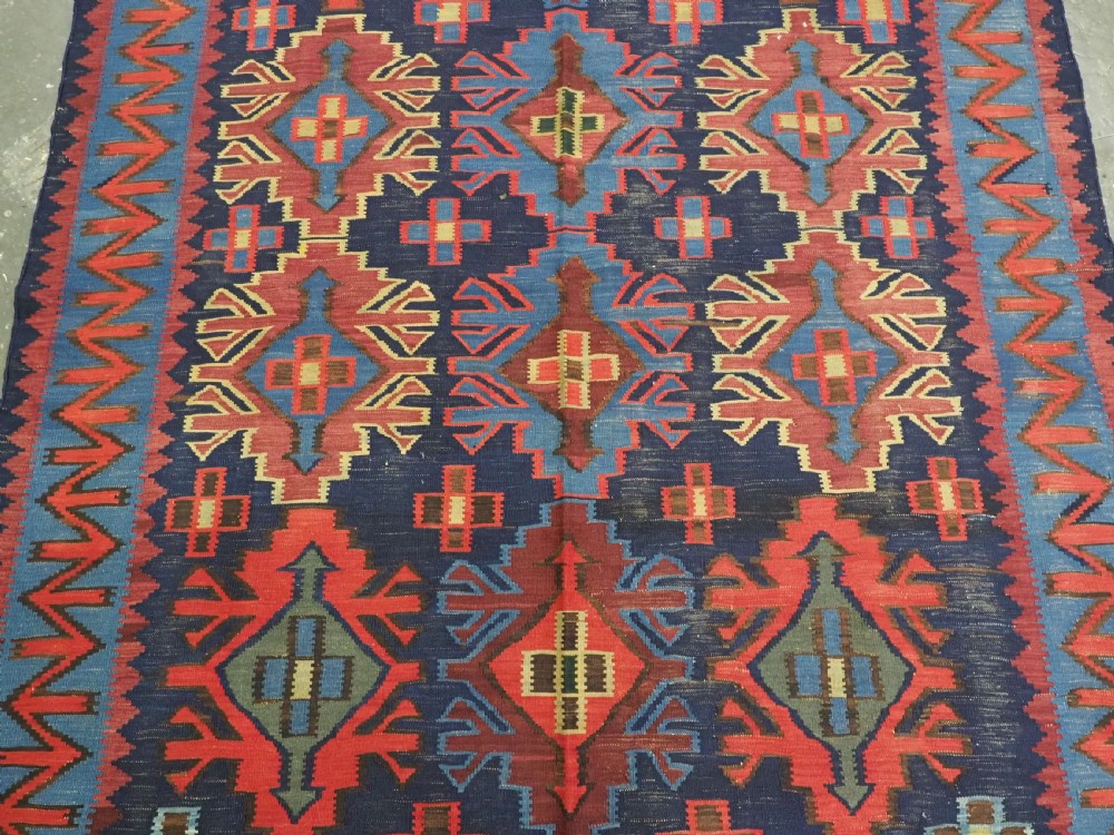 An antique flatweave Caucasian Kuba kilim rug featuring geometric shield medallions in red, blue, and cream across a dark navy field. View IV