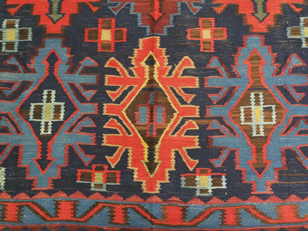 An antique flatweave Caucasian Kuba kilim rug featuring geometric shield medallions in red, blue, and cream across a dark navy field. View X