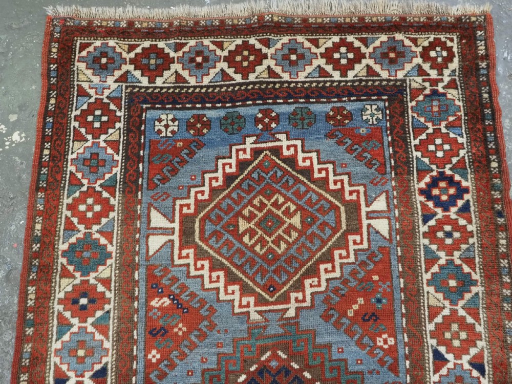 An antique hand-knotted wool rug featuring three hooked diamond medallions along a central blue field surrounded by geometric patterned borders in terracotta, red, and cream. View III