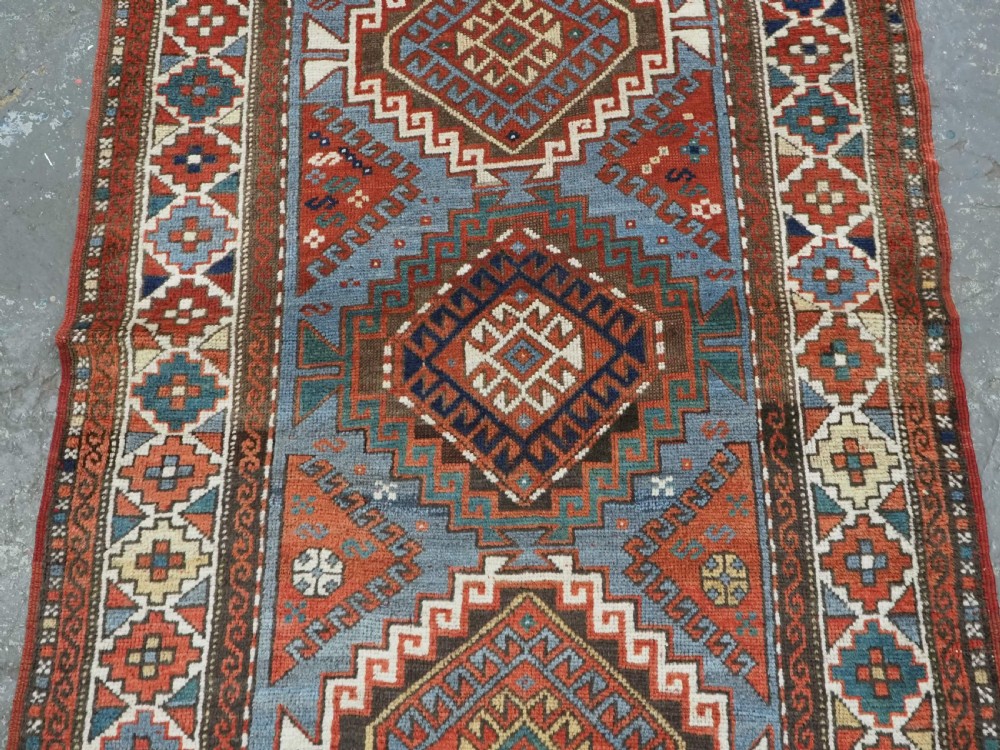 An antique hand-knotted wool rug featuring three hooked diamond medallions along a central blue field surrounded by geometric patterned borders in terracotta, red, and cream. View IV