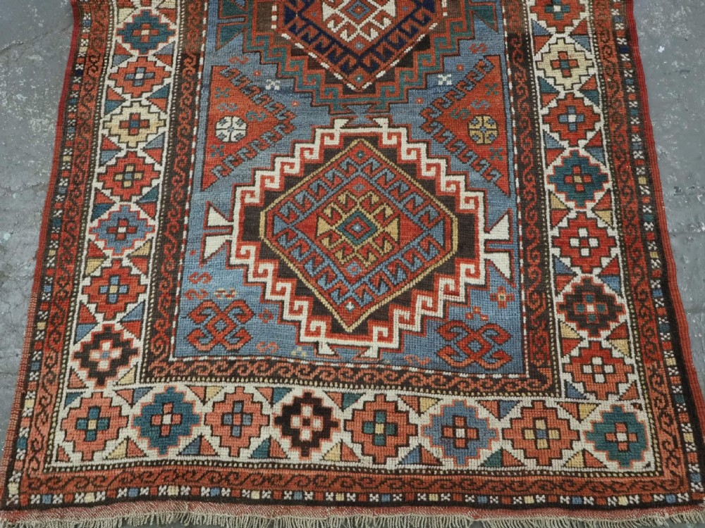 An antique hand-knotted wool rug featuring three hooked diamond medallions along a central blue field surrounded by geometric patterned borders in terracotta, red, and cream. View V