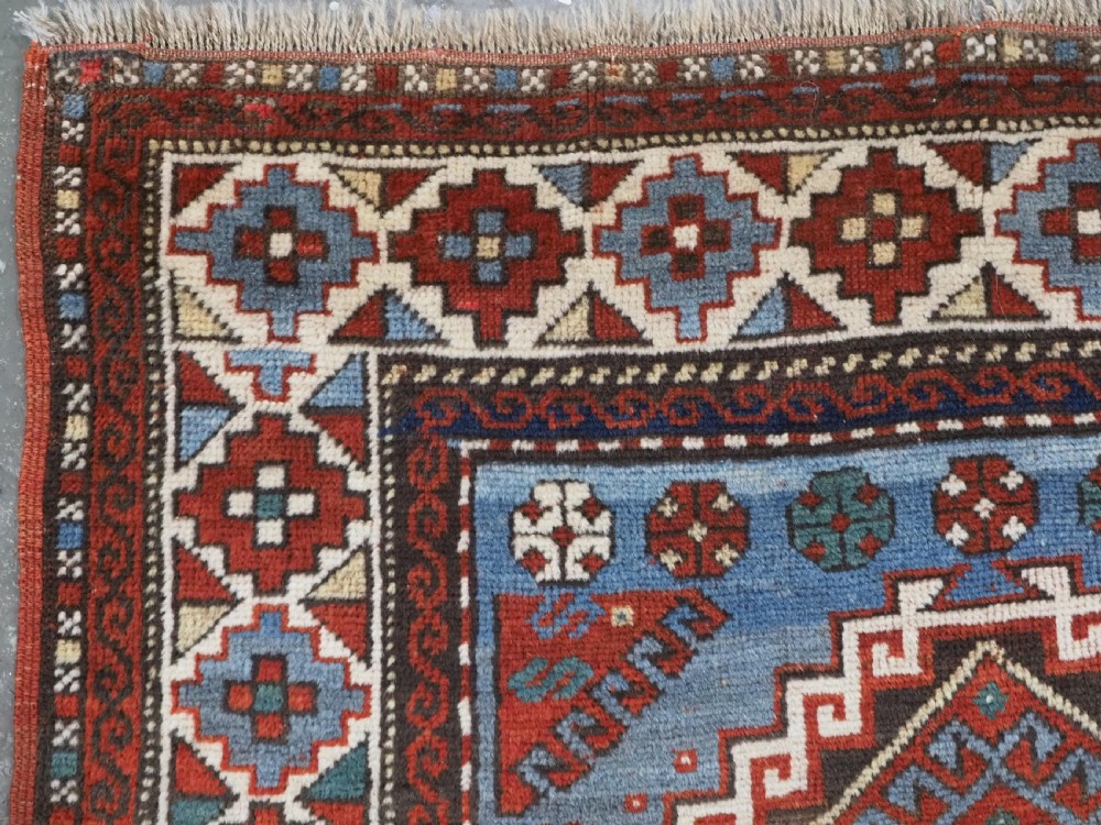 An antique hand-knotted wool rug featuring three hooked diamond medallions along a central blue field surrounded by geometric patterned borders in terracotta, red, and cream. View VI