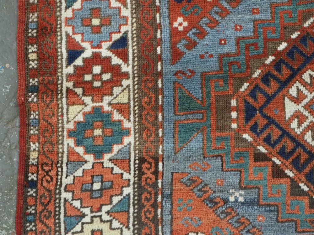 An antique hand-knotted wool rug featuring three hooked diamond medallions along a central blue field surrounded by geometric patterned borders in terracotta, red, and cream. View VII