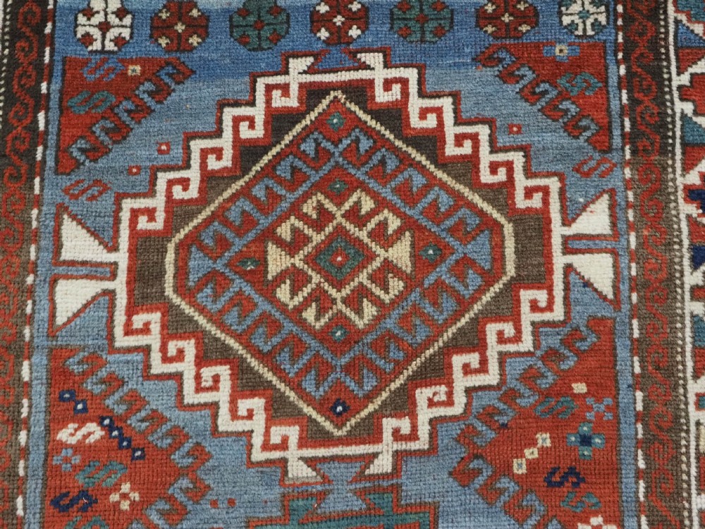 An antique hand-knotted wool rug featuring three hooked diamond medallions along a central blue field surrounded by geometric patterned borders in terracotta, red, and cream. View VIII