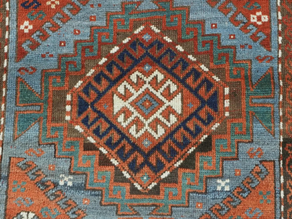 An antique hand-knotted wool rug featuring three hooked diamond medallions along a central blue field surrounded by geometric patterned borders in terracotta, red, and cream. View IX