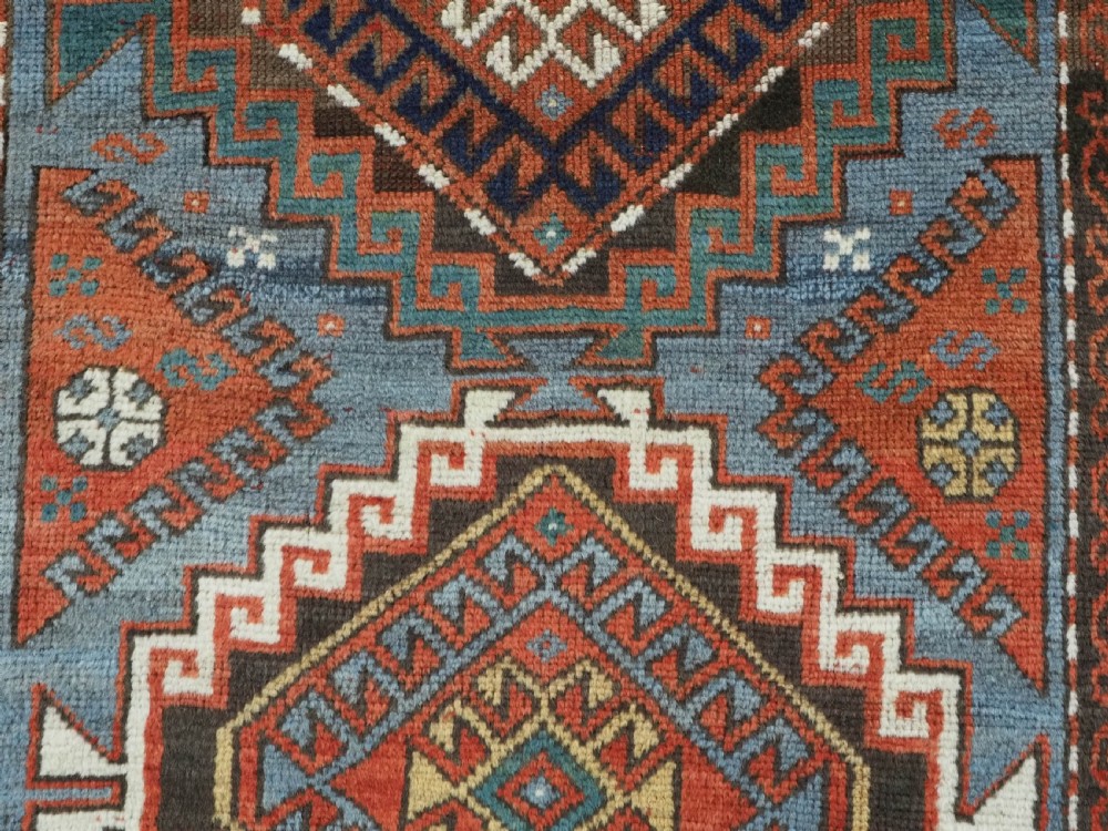 An antique hand-knotted wool rug featuring three hooked diamond medallions along a central blue field surrounded by geometric patterned borders in terracotta, red, and cream. View X