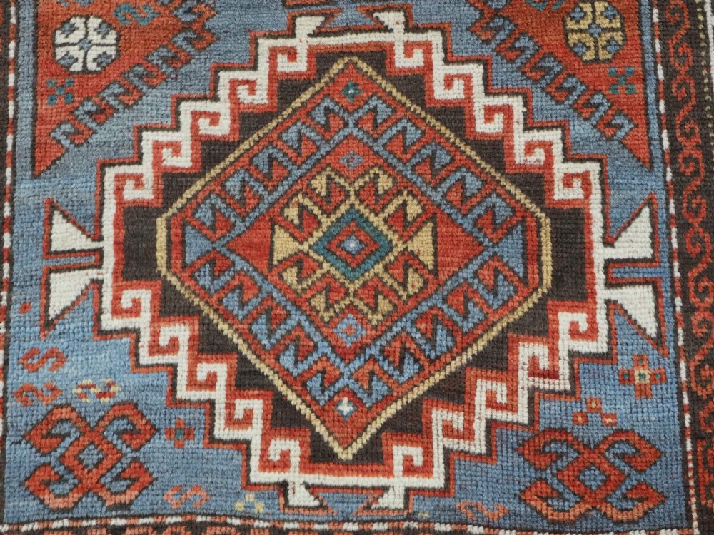 An antique hand-knotted wool rug featuring three hooked diamond medallions along a central blue field surrounded by geometric patterned borders in terracotta, red, and cream. View XI