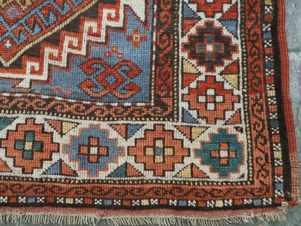 An antique hand-knotted wool rug featuring three hooked diamond medallions along a central blue field surrounded by geometric patterned borders in terracotta, red, and cream. View XII