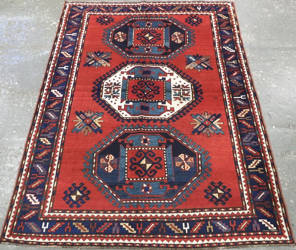 An antique Caucasian Kazak hand-knotted wool rug featuring three large geometric medallions along a rich red field surrounded by a dark blue border.