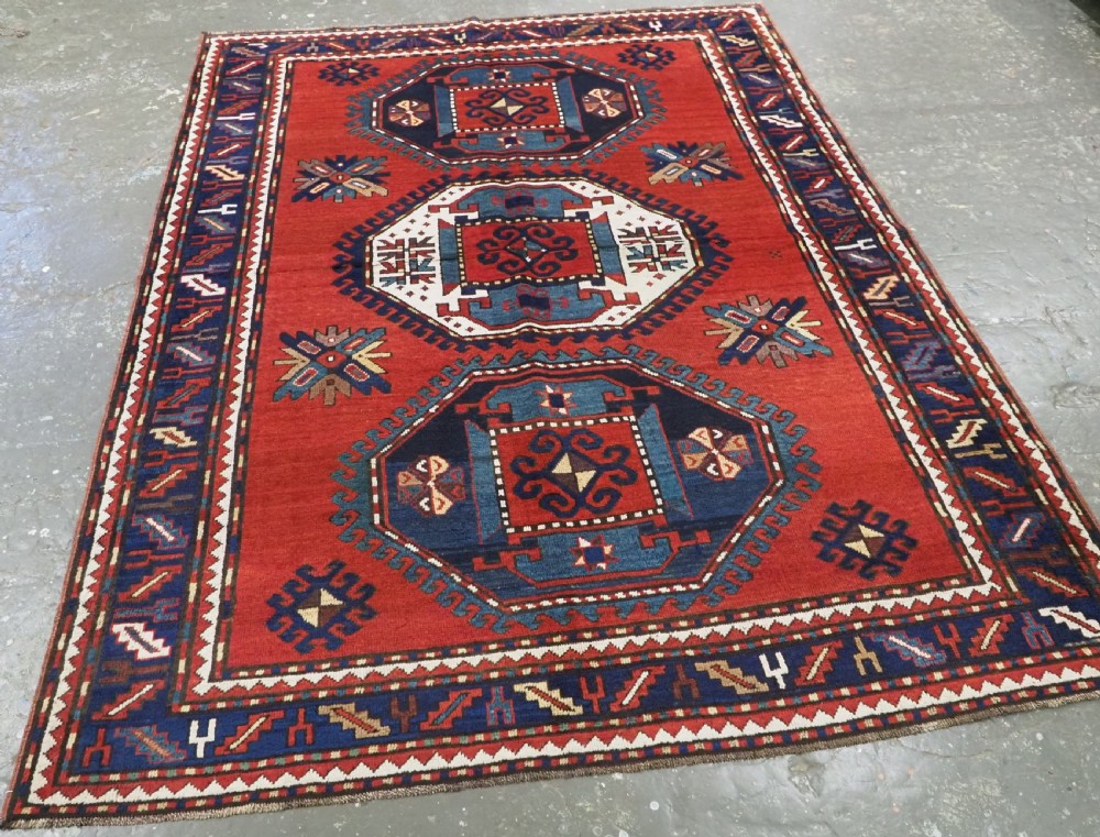 An antique Caucasian Kazak hand-knotted wool rug featuring three large geometric medallions along a rich red field surrounded by a dark blue border. View II
