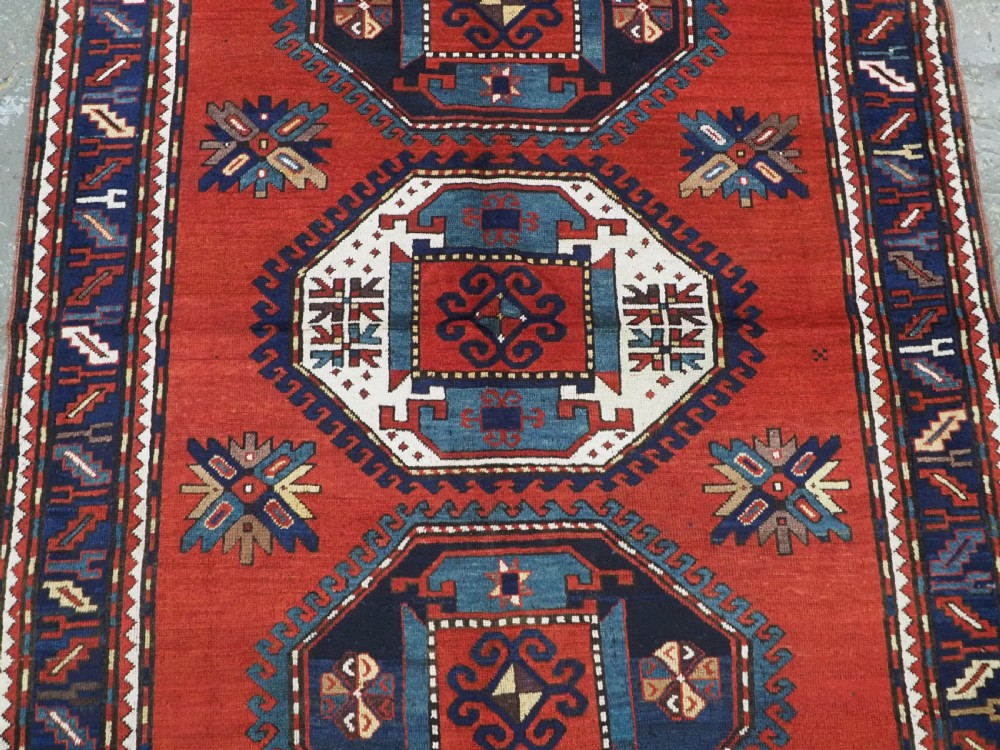 An antique Caucasian Kazak hand-knotted wool rug featuring three large geometric medallions along a rich red field surrounded by a dark blue border. View IV