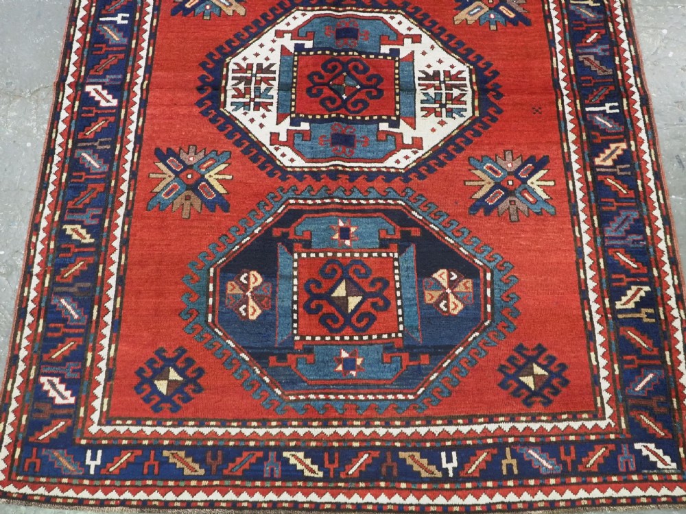 An antique Caucasian Kazak hand-knotted wool rug featuring three large geometric medallions along a rich red field surrounded by a dark blue border. View V