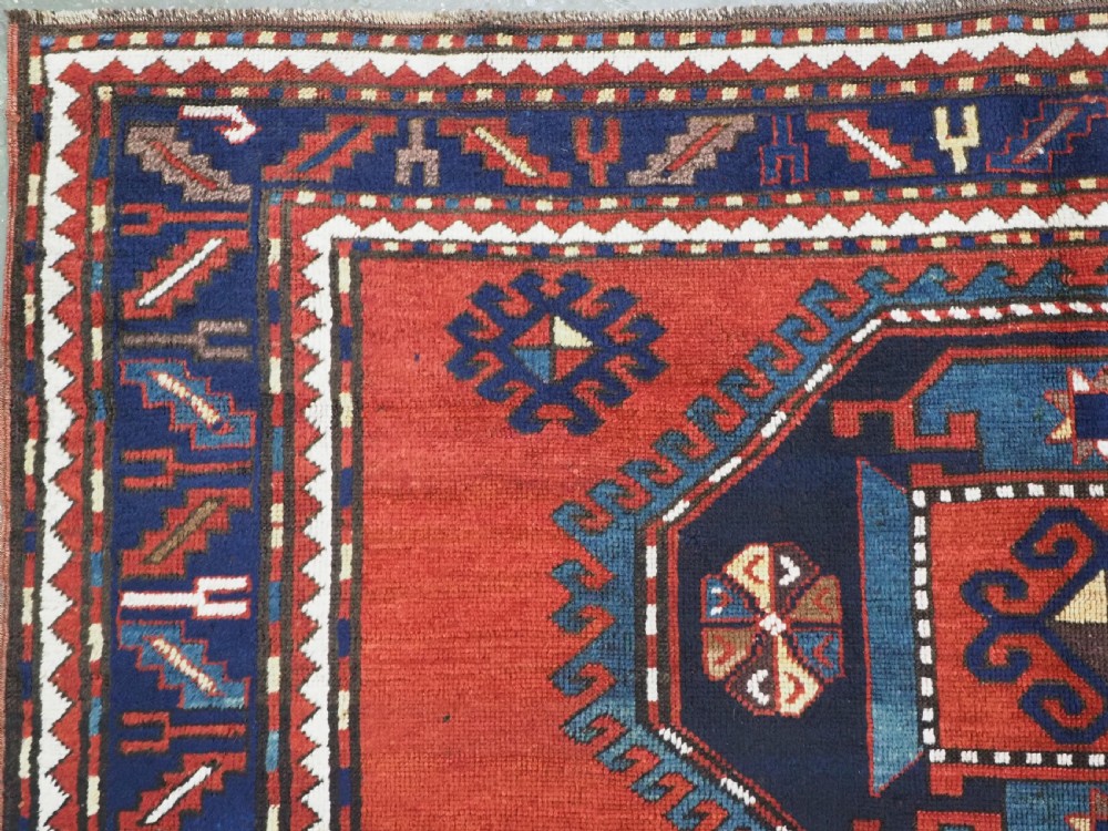 An antique Caucasian Kazak hand-knotted wool rug featuring three large geometric medallions along a rich red field surrounded by a dark blue border. View VI