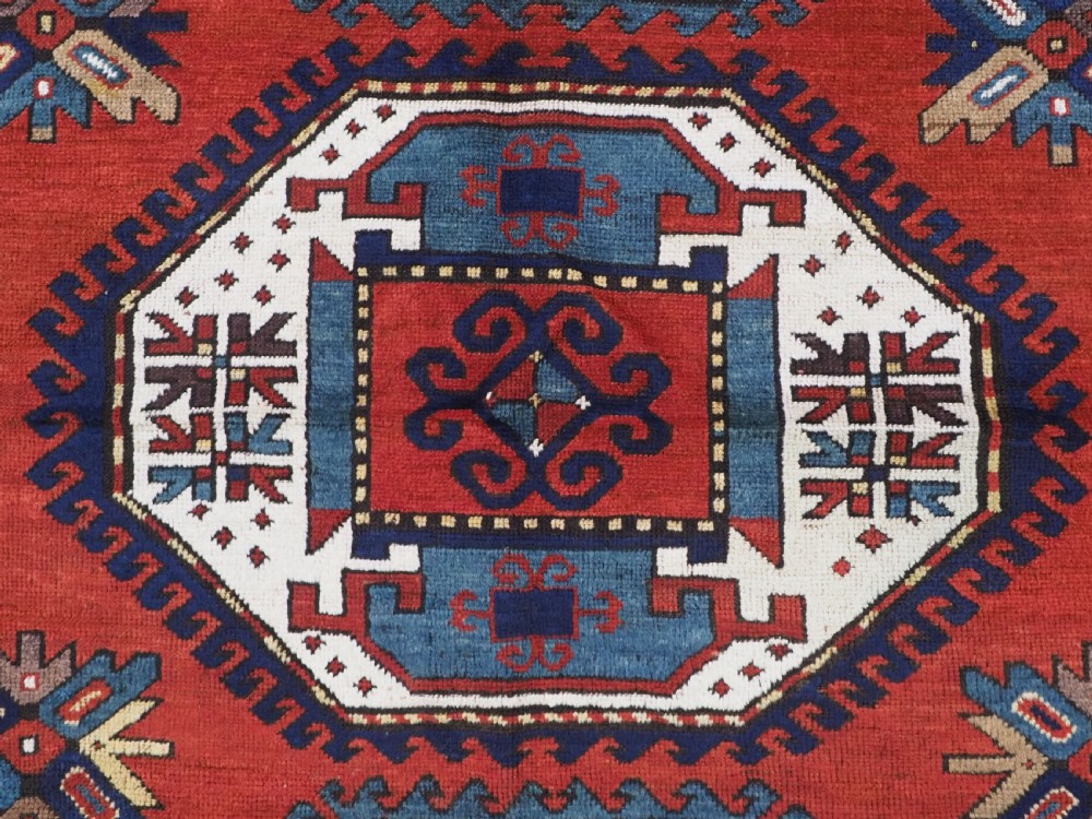An antique Caucasian Kazak hand-knotted wool rug featuring three large geometric medallions along a rich red field surrounded by a dark blue border. View X