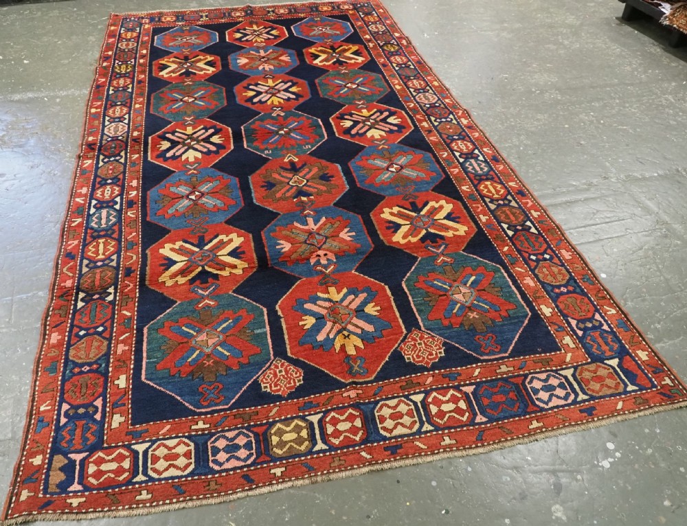 An antique hand-knotted wool rug featuring columns of red and blue hexagonal medallions on a deep navy field framed by a geometric multi-band border. View II