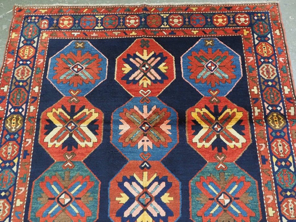 An antique hand-knotted wool rug featuring columns of red and blue hexagonal medallions on a deep navy field framed by a geometric multi-band border. View III
