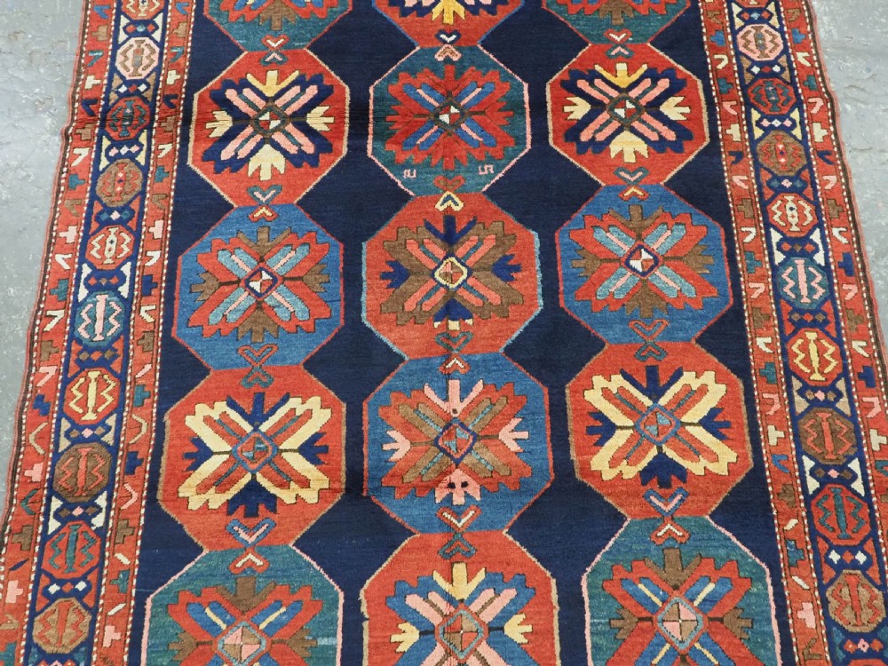 An antique hand-knotted wool rug featuring columns of red and blue hexagonal medallions on a deep navy field framed by a geometric multi-band border. View IV