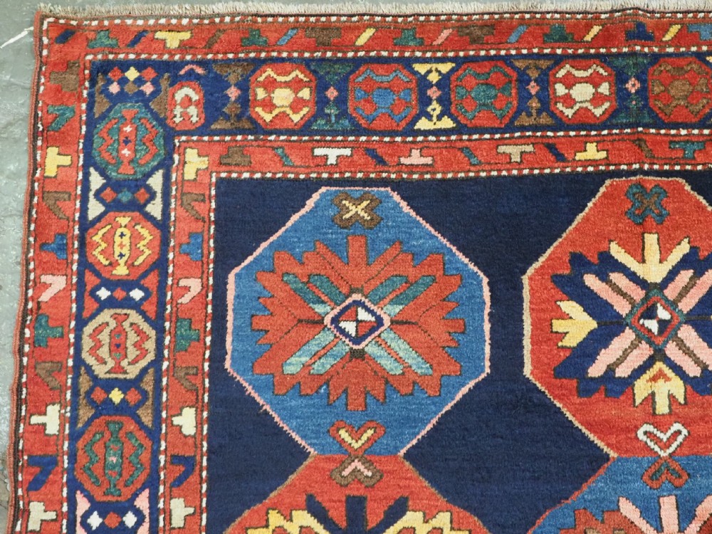 An antique hand-knotted wool rug featuring columns of red and blue hexagonal medallions on a deep navy field framed by a geometric multi-band border. View V
