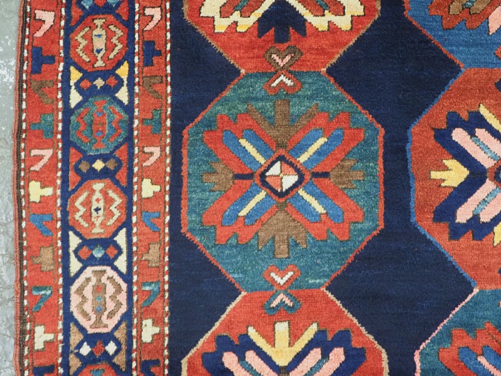 An antique hand-knotted wool rug featuring columns of red and blue hexagonal medallions on a deep navy field framed by a geometric multi-band border. View VI